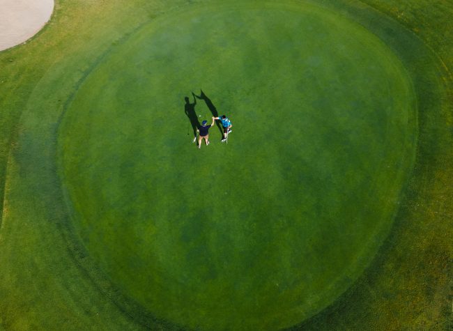 Two golfers are playing on a vibrant green golf course with a sand bunker and surrounding trees under clear skies